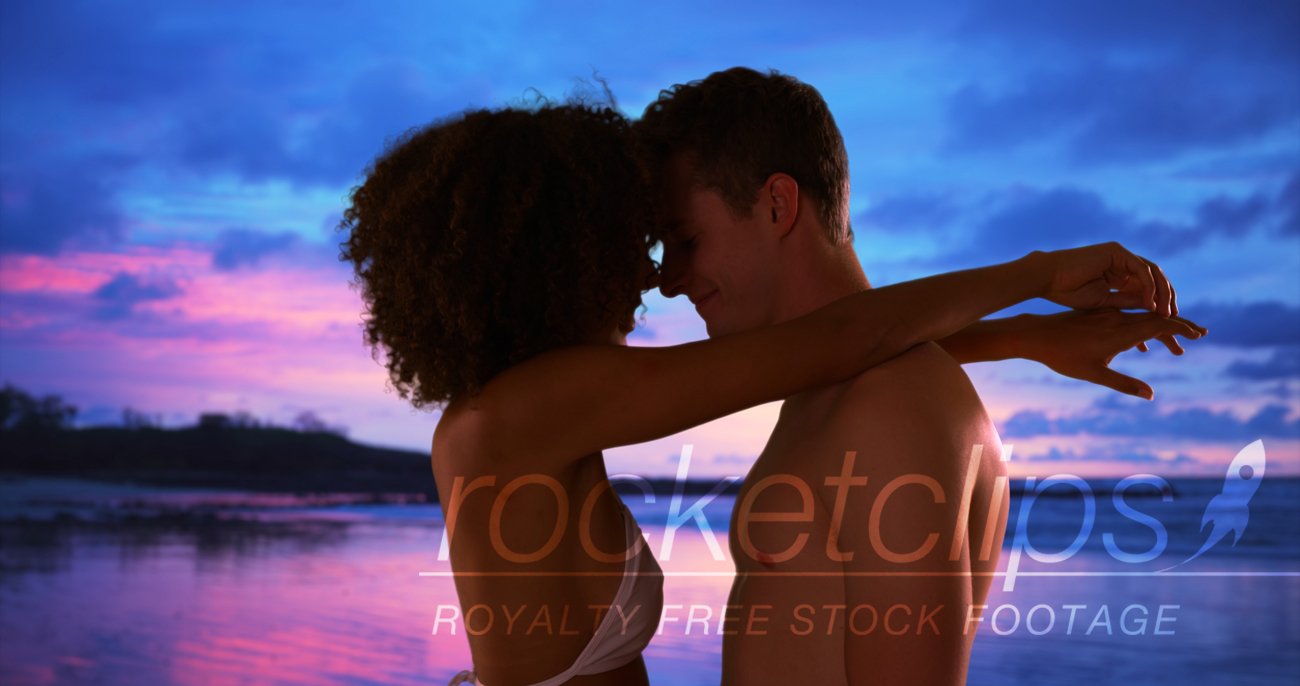 African woman with arms around boyfriend under pink and blue sky.