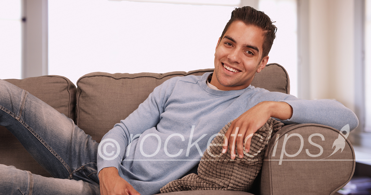 Portrait of handsome young Hispanic man sitting on couch in living room smiling at camera