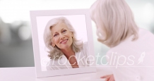 Lovely Caucasian senior lady looking at herself in mirror in clean white setting