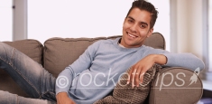 Portrait of handsome young Hispanic man sitting on couch in living room smiling at camera